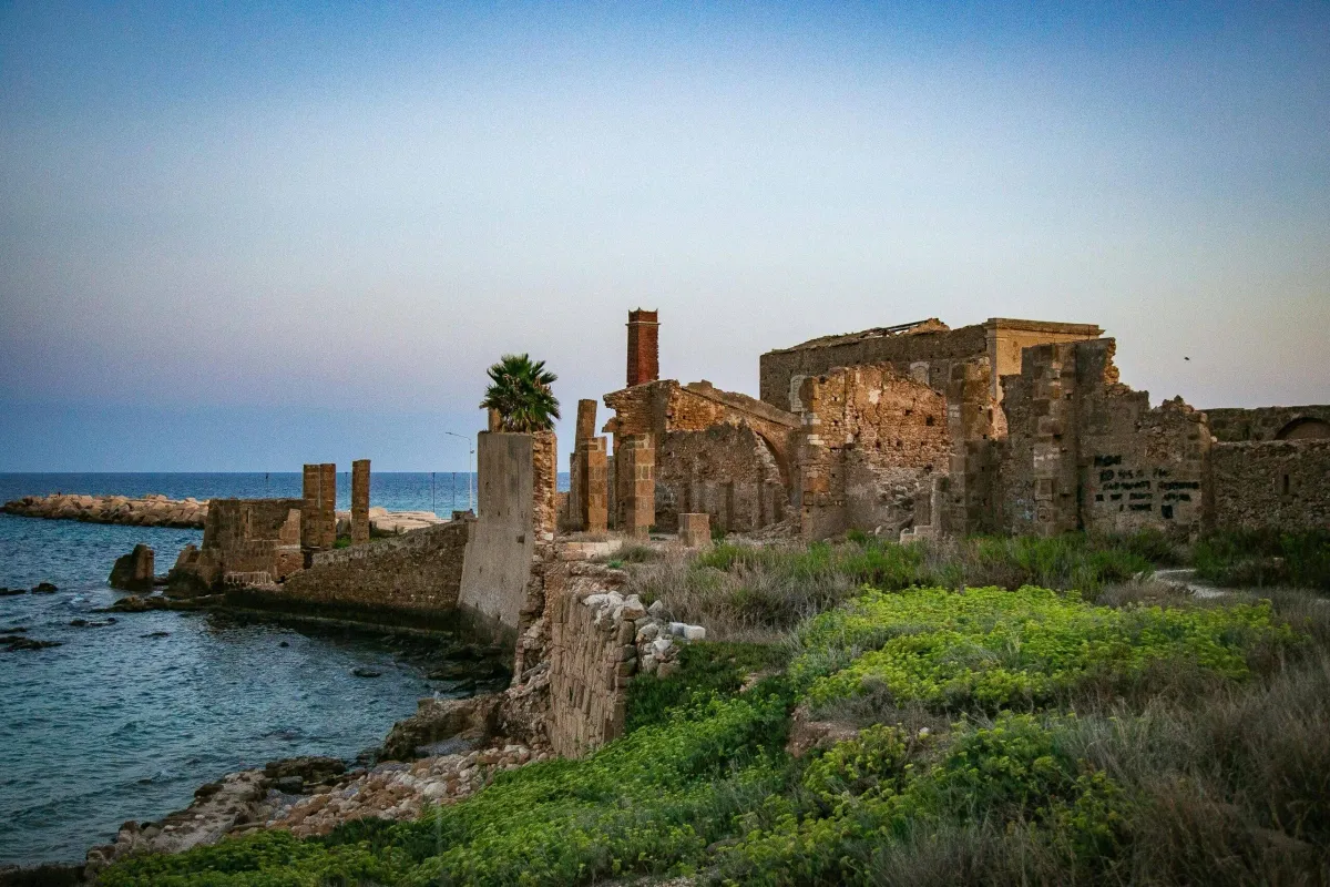 I resti suggestivi della Vecchia Tonnara di Avola in Sicilia, illuminati dalla luce soffusa del crepuscolo. Le antiche mura in pietra e una ciminiera diroccata si ergono tra la vegetazione spontanea verdeggiante, affacciandosi direttamente su una scogliera bagnata dal mare calmo.